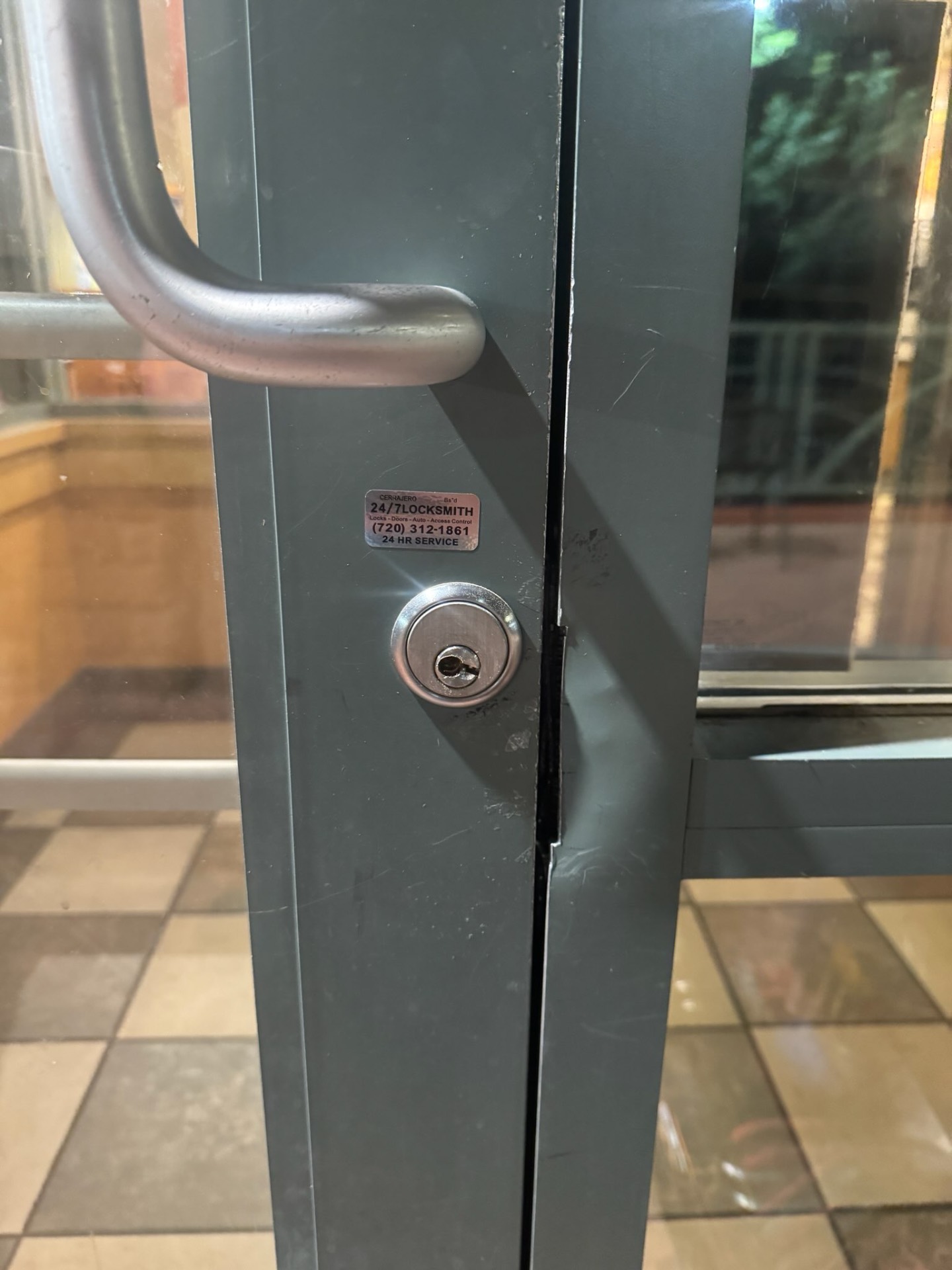Close-up of a dark metal-framed glass commercial door with a large pull handle, round keyed cylinder, scuffed frame, and locksmith sticker.