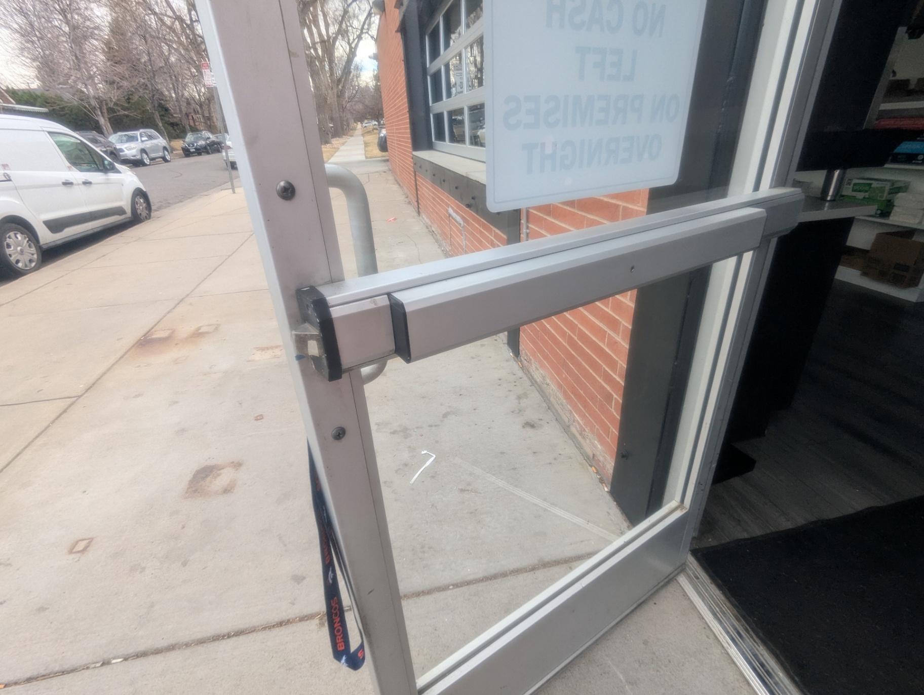 Locksmith handling a rim-cylinder component and keys at a Denver commercial glass storefront entrance with panic-bar hardware visible nearby. - view 2