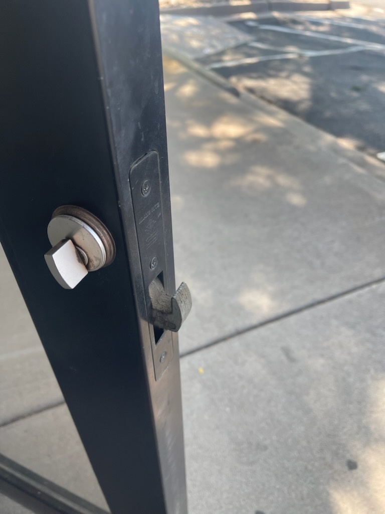 Black-framed glass commercial storefront door with a round exterior cylinder, vertical pull handle, interior thumbturn, and exposed mortise latch hardware - view 3