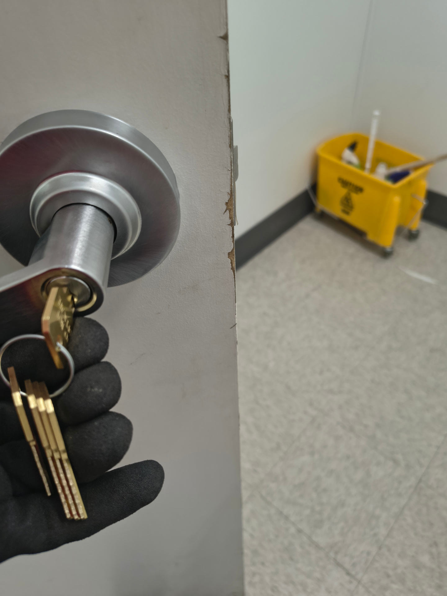 Gloved locksmith holding a gold-colored cylinder and keys beside a silver commercial restroom door knob - view 2