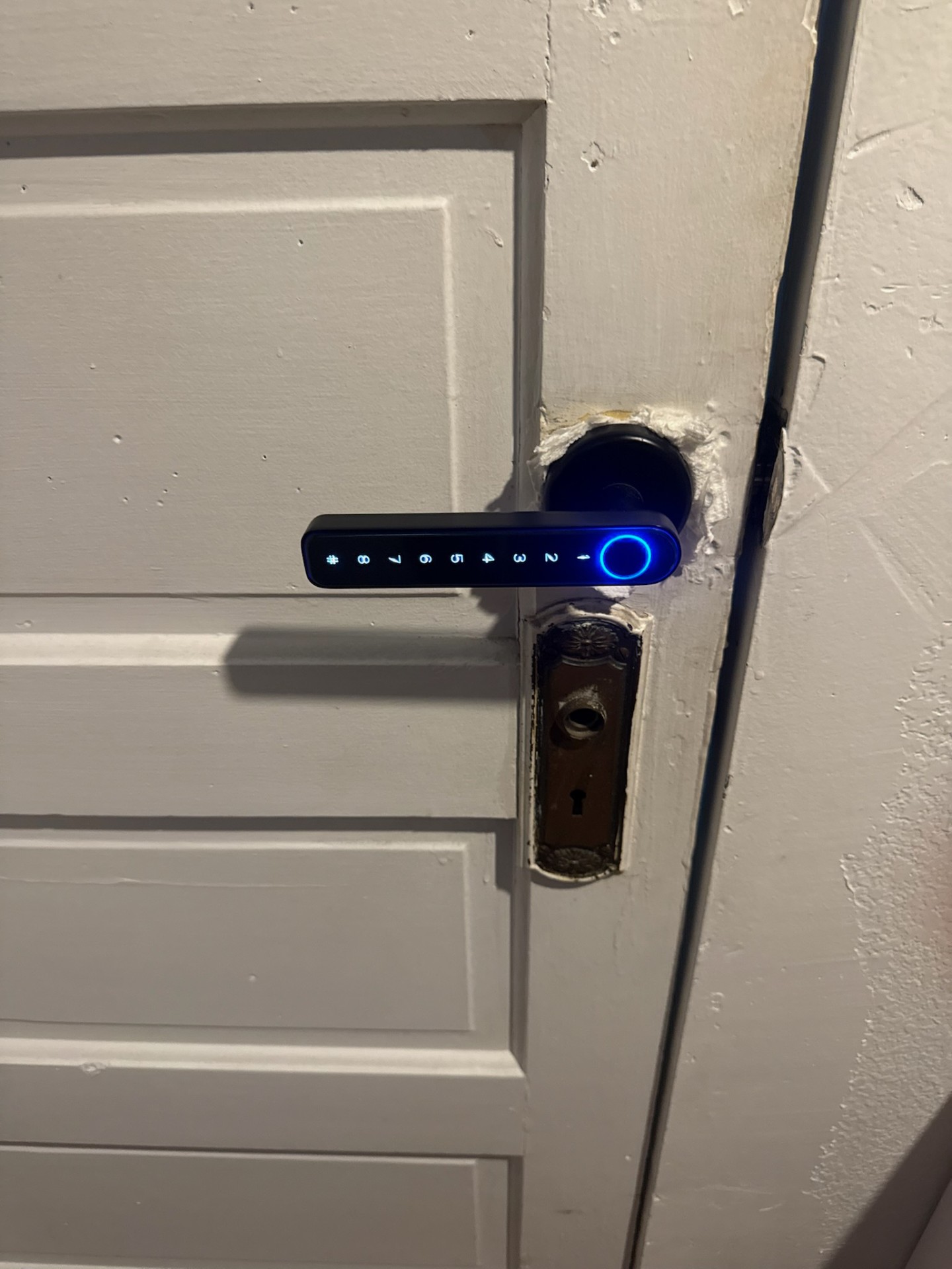 Close-up of a black blue-illuminated electronic keypad lever lock above separate lower lock hardware on a white paneled door in Boulder