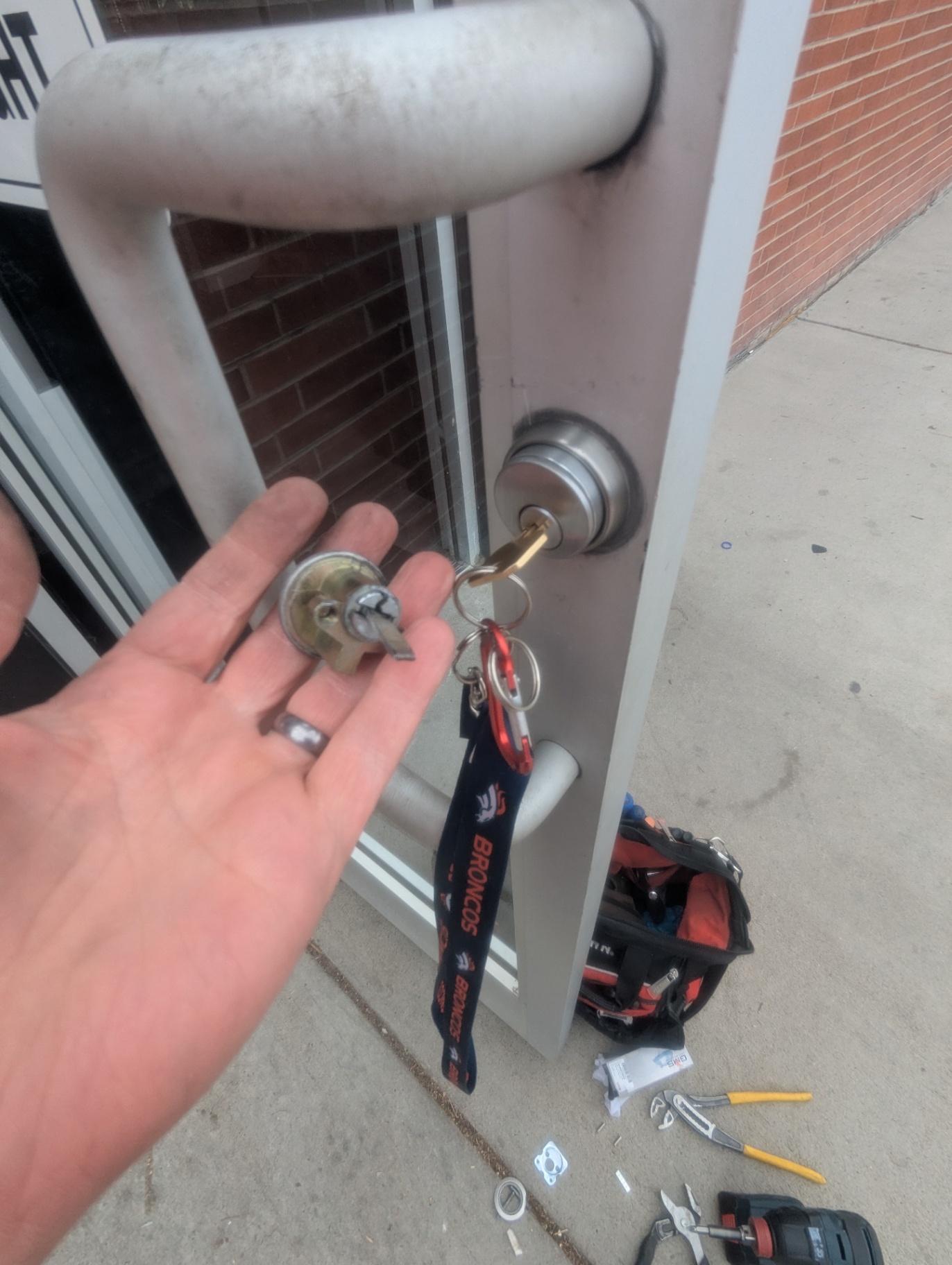 Locksmith handling a rim-cylinder component and keys at a Denver commercial glass storefront entrance with panic-bar hardware visible nearby. - view 3