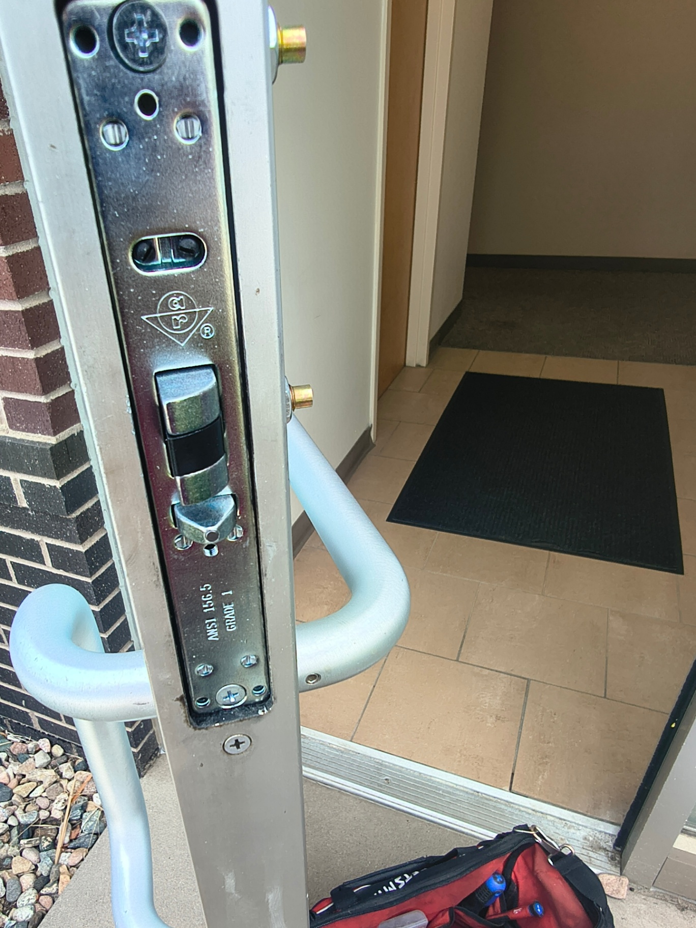 Open glass-and-aluminum commercial door showing exposed mortise lock hardware, pull handle, and black push exit device