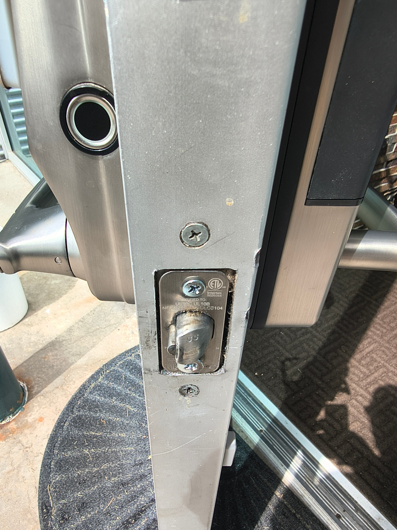 Close-up of exposed commercial door-edge latch hardware, lock cylinders, mounting marks, and an electronic-looking lock at a Lakewood business entrance. - view 3