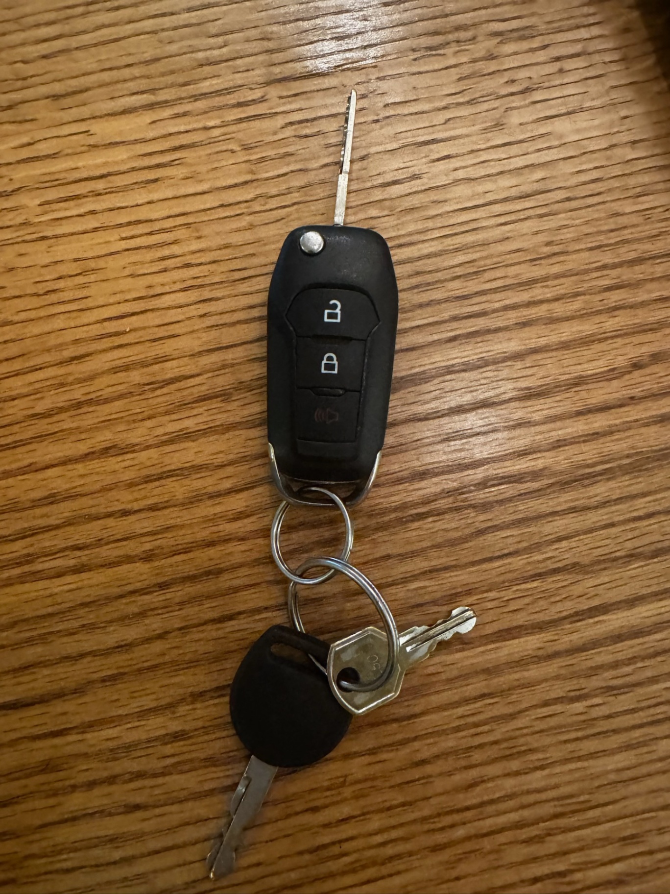 Black automotive remote key fob with lock and unlock buttons, an extended metal blade, and additional keys on a metal ring on a wooden surface.