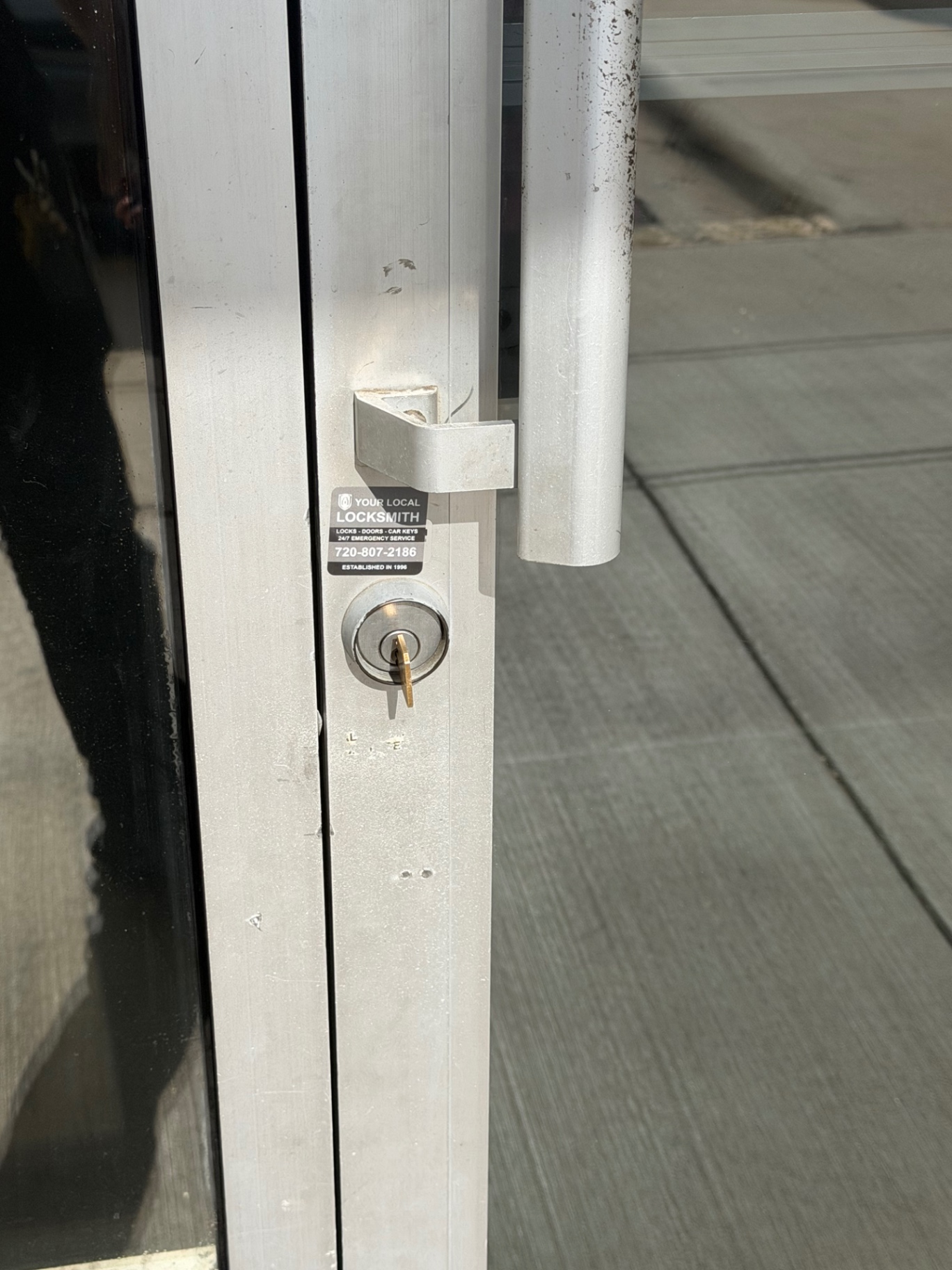 Close-up of a worn keyed cylindrical lock and pull handle on an aluminum-framed glass storefront door in Denver. - view 2