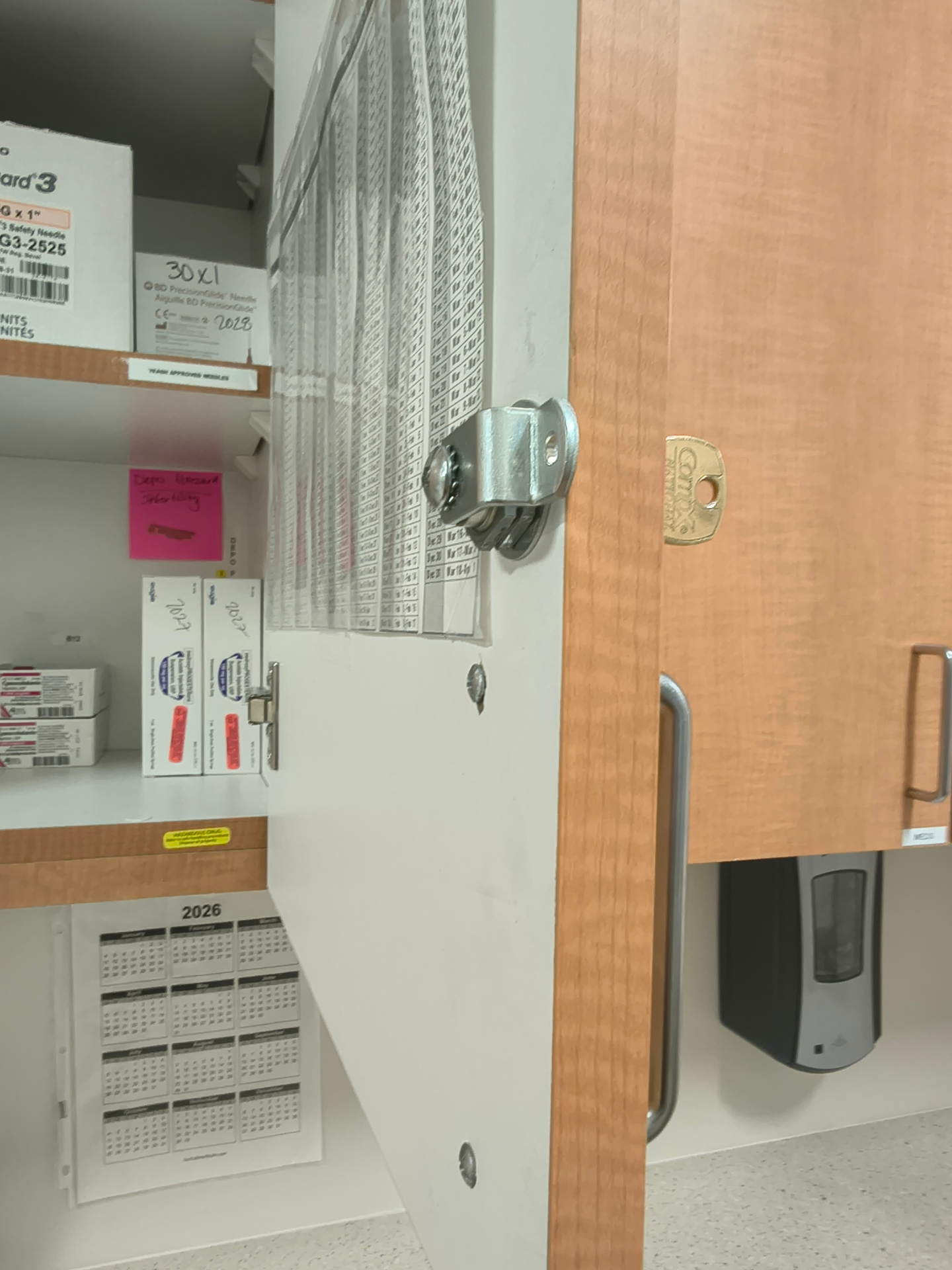 Exposed cylindrical cam lock with brass-colored key on a wood-finish interior cupboard beside an electronic access device in Westminster
