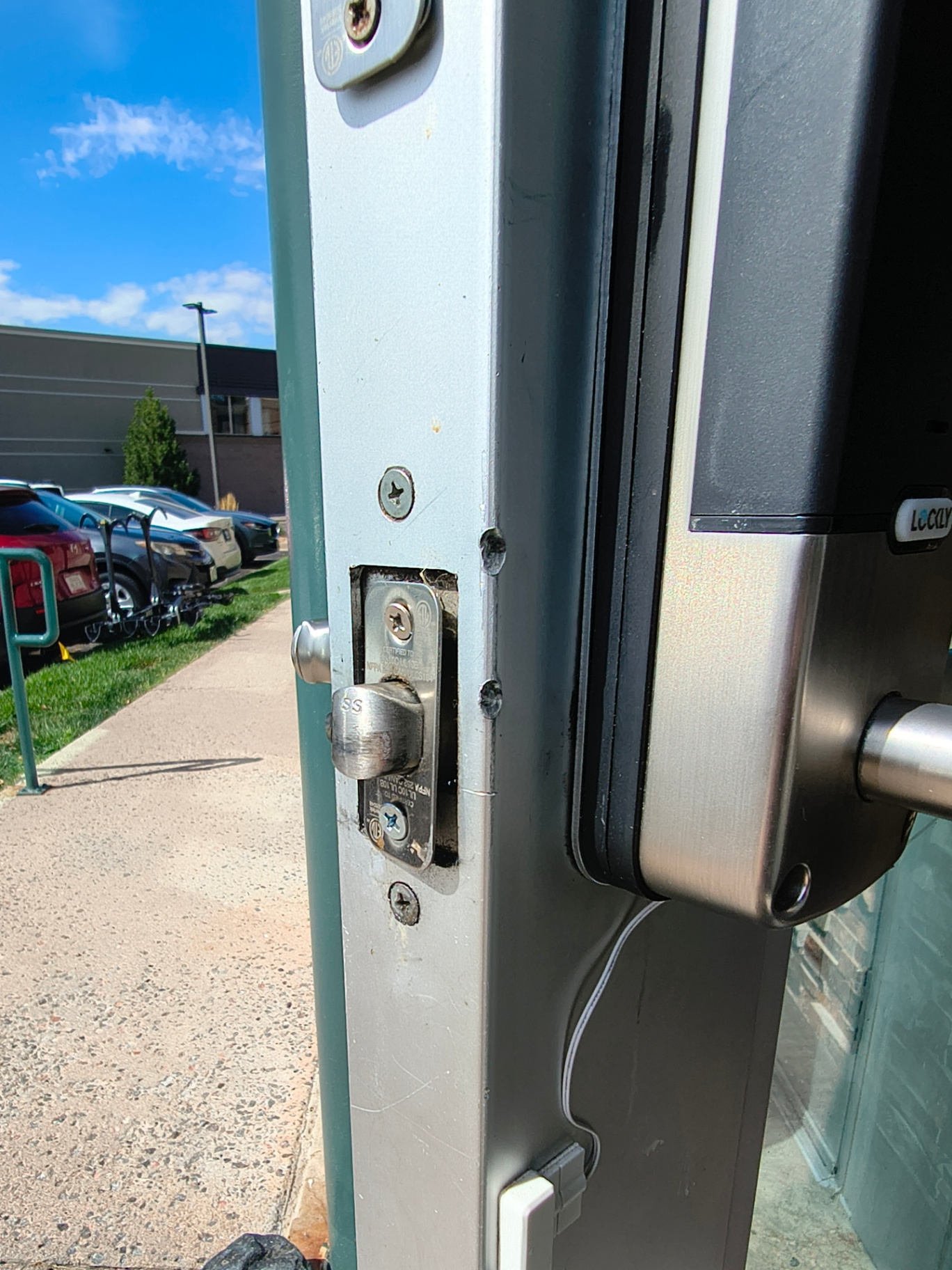 Close-up of exposed commercial door-edge latch hardware, lock cylinders, mounting marks, and an electronic-looking lock at a Lakewood business entrance. - view 2