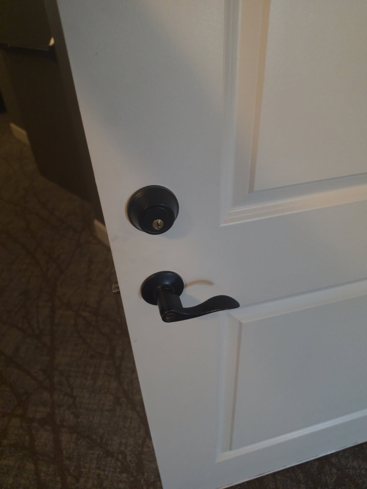 White paneled interior door in Littleton with a black deadbolt above a black lever handle, shown partially open. - view 2