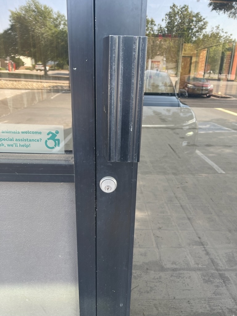 Black-framed glass commercial storefront door with a round exterior cylinder, vertical pull handle, interior thumbturn, and exposed mortise latch hardware