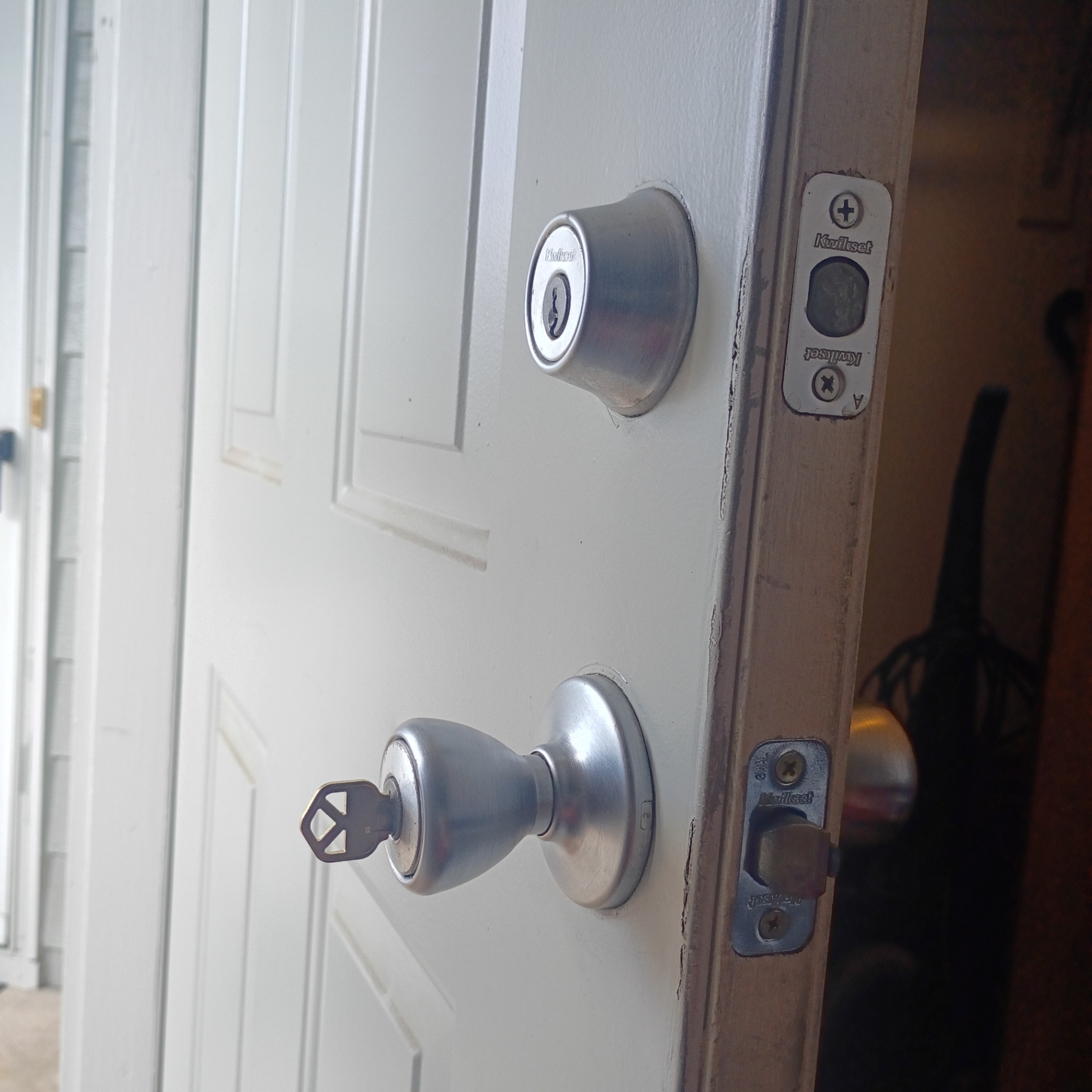 White paneled residential door partially open with a silver keyed deadbolt above a silver keyed entry knob, visible latch and strike plates, and a key inserted in the lower lock.