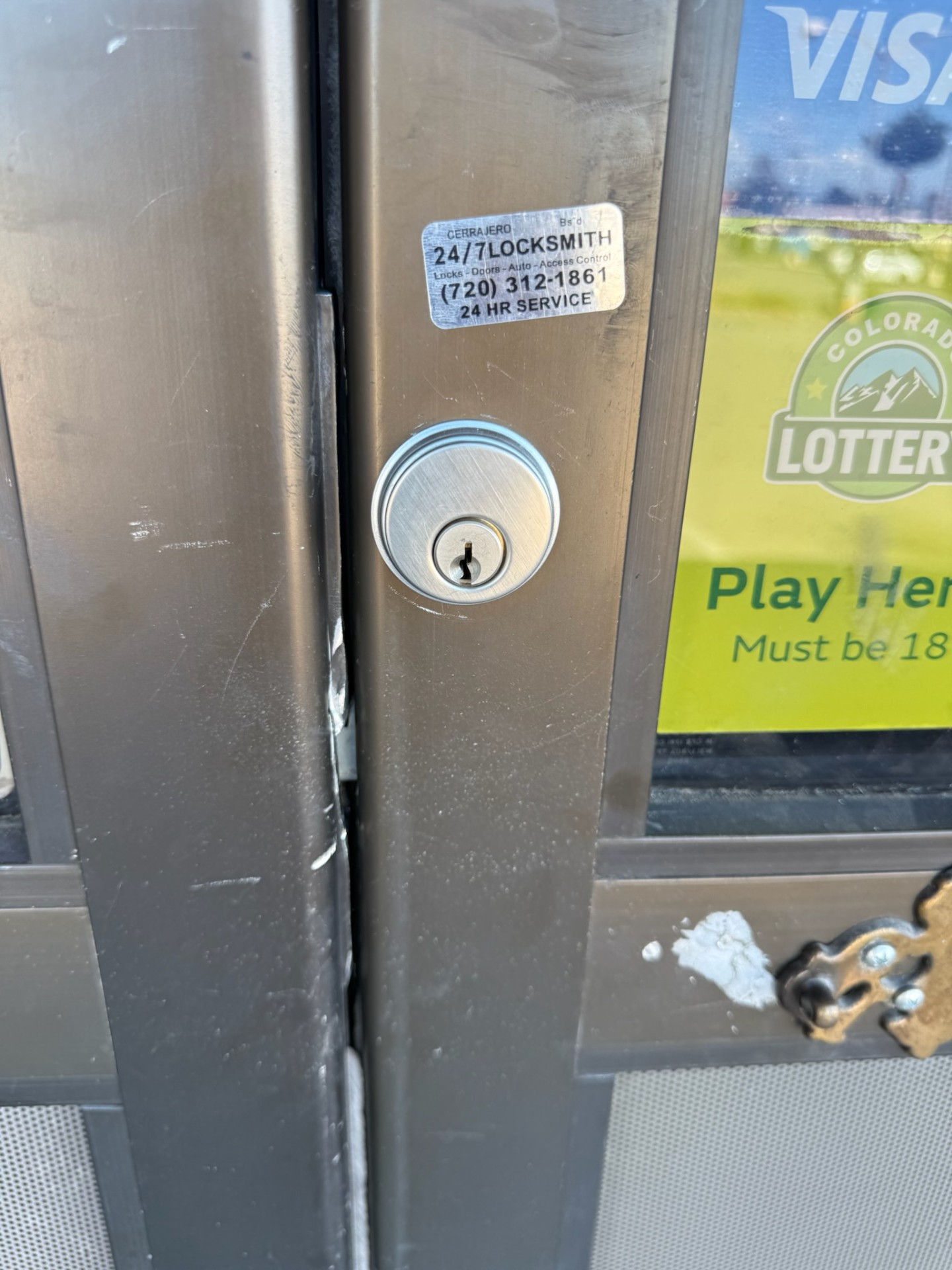 Close-up of a round keyed cylinder on a scratched metal-framed glass commercial door beside lottery signage