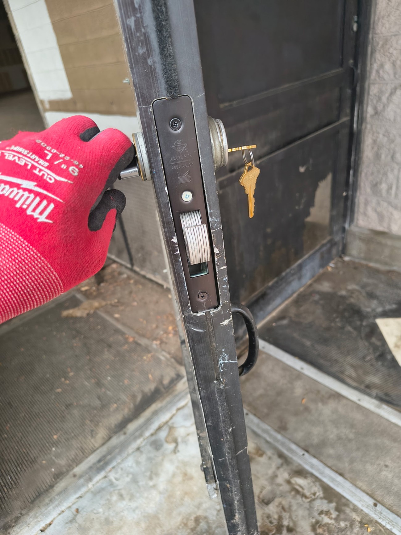 Gloved locksmith holds a removed mortise lock assembly beside a commercial door with a keyed cylinder and keys visible. - view 2