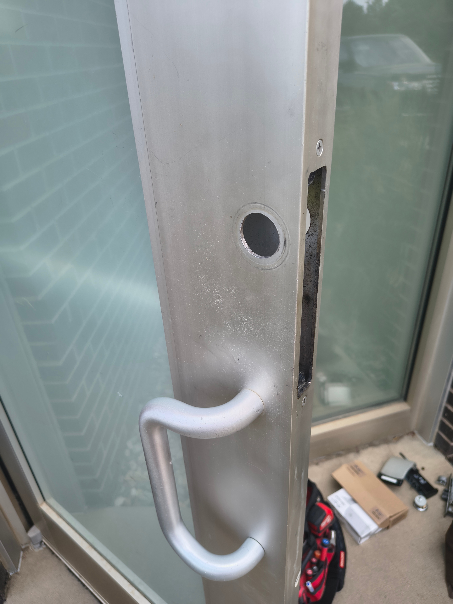 Open glass-and-aluminum commercial door showing exposed mortise lock hardware, pull handle, and black push exit device - view 2