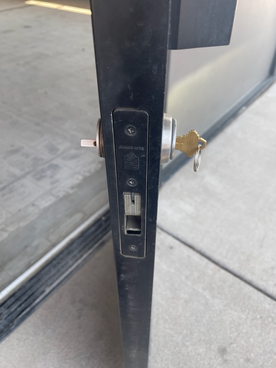 Black-framed glass commercial storefront door with a round exterior cylinder, vertical pull handle, interior thumbturn, and exposed mortise latch hardware - view 4