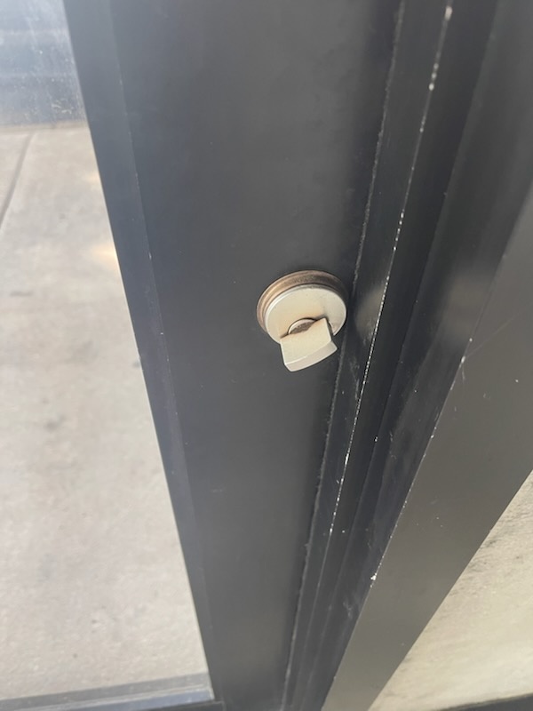Black-framed glass commercial storefront door with a round exterior cylinder, vertical pull handle, interior thumbturn, and exposed mortise latch hardware - view 2