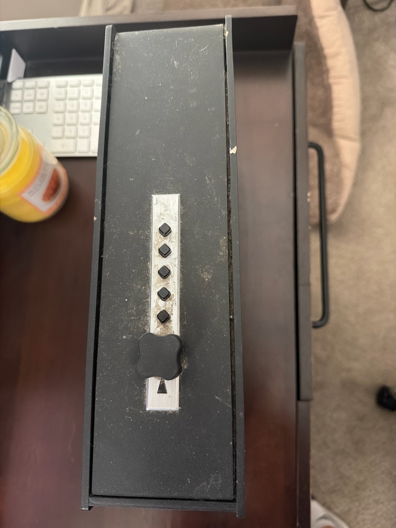 Black rectangular gun safe on a desk in an office workspace, with a vertical row of push buttons, a large turning handle, and a dusty scuffed surface.