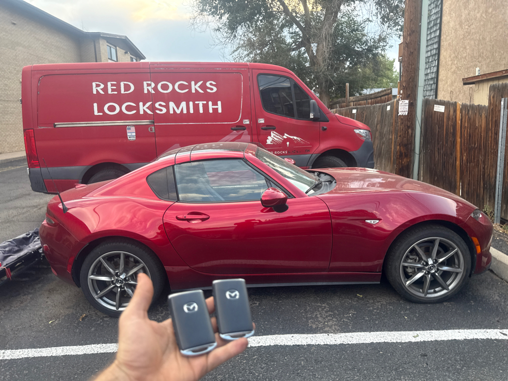 Two Mazda-logo key fobs held in front of a red sports coupe with a red locksmith van parked behind it in Denver