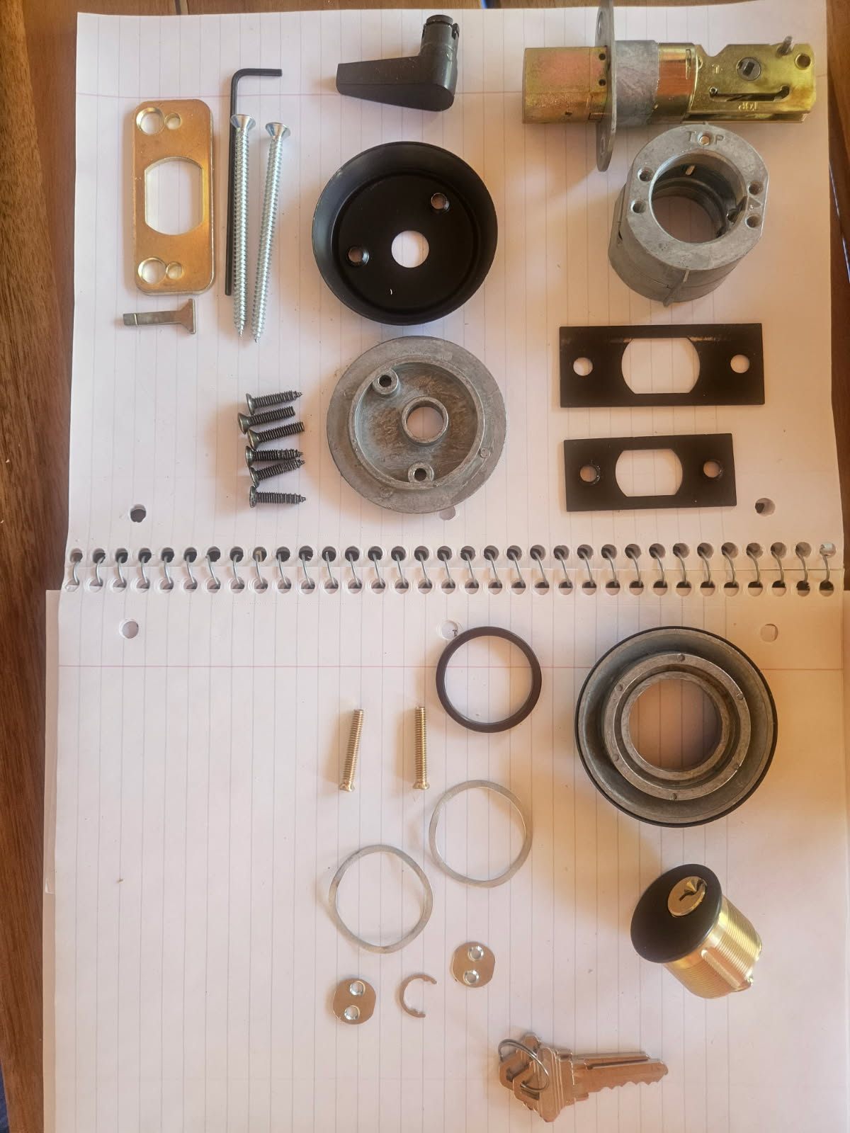 Separated cylindrical door lock components, mounting plates, screws, retaining rings, and keys arranged on paper for a Lakewood lock change.