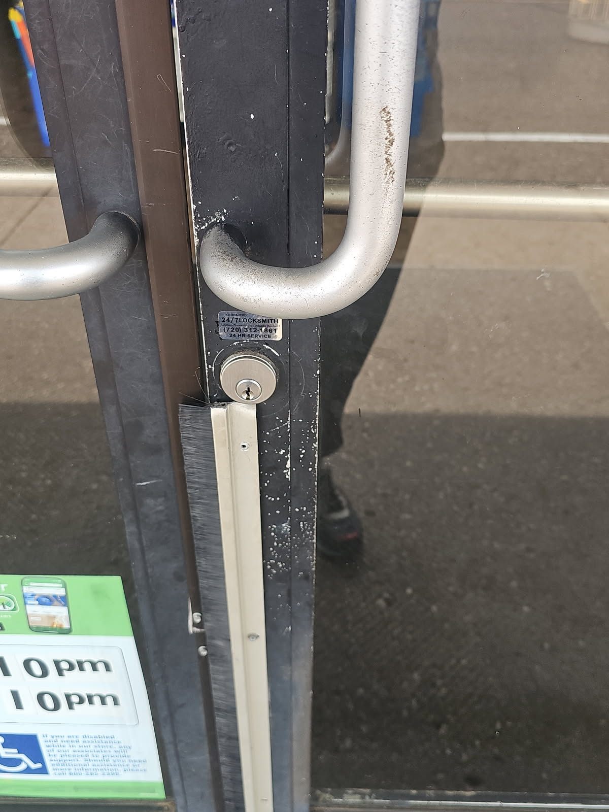 Close-up of a keyed cylinder below a large vertical pull handle on a worn black-framed glass storefront door in Lakewood, Colorado