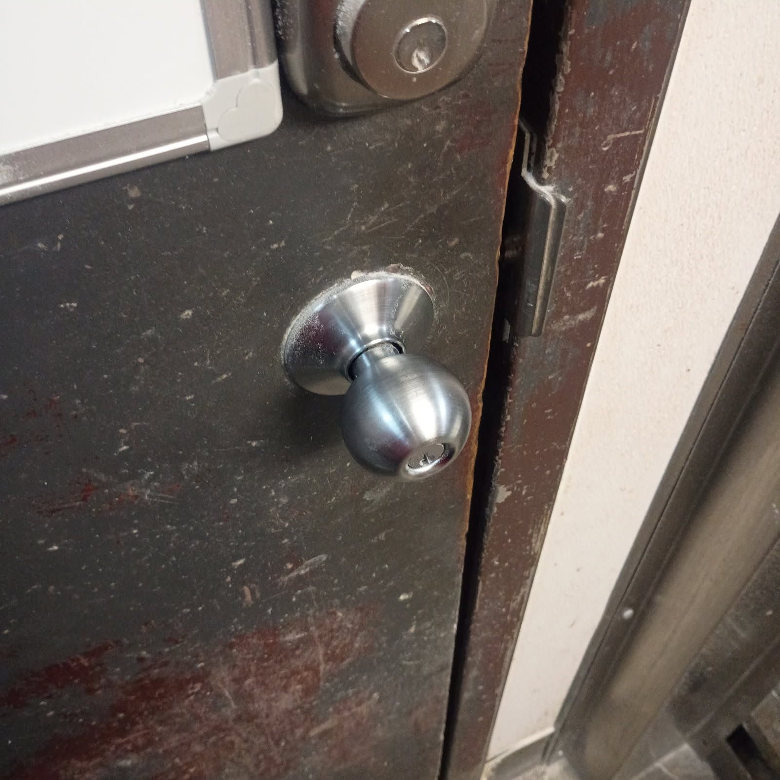 Close-up of a worn dark commercial door with a silver keyed deadbolt and round keyed knob beside a chipped, misaligned frame.