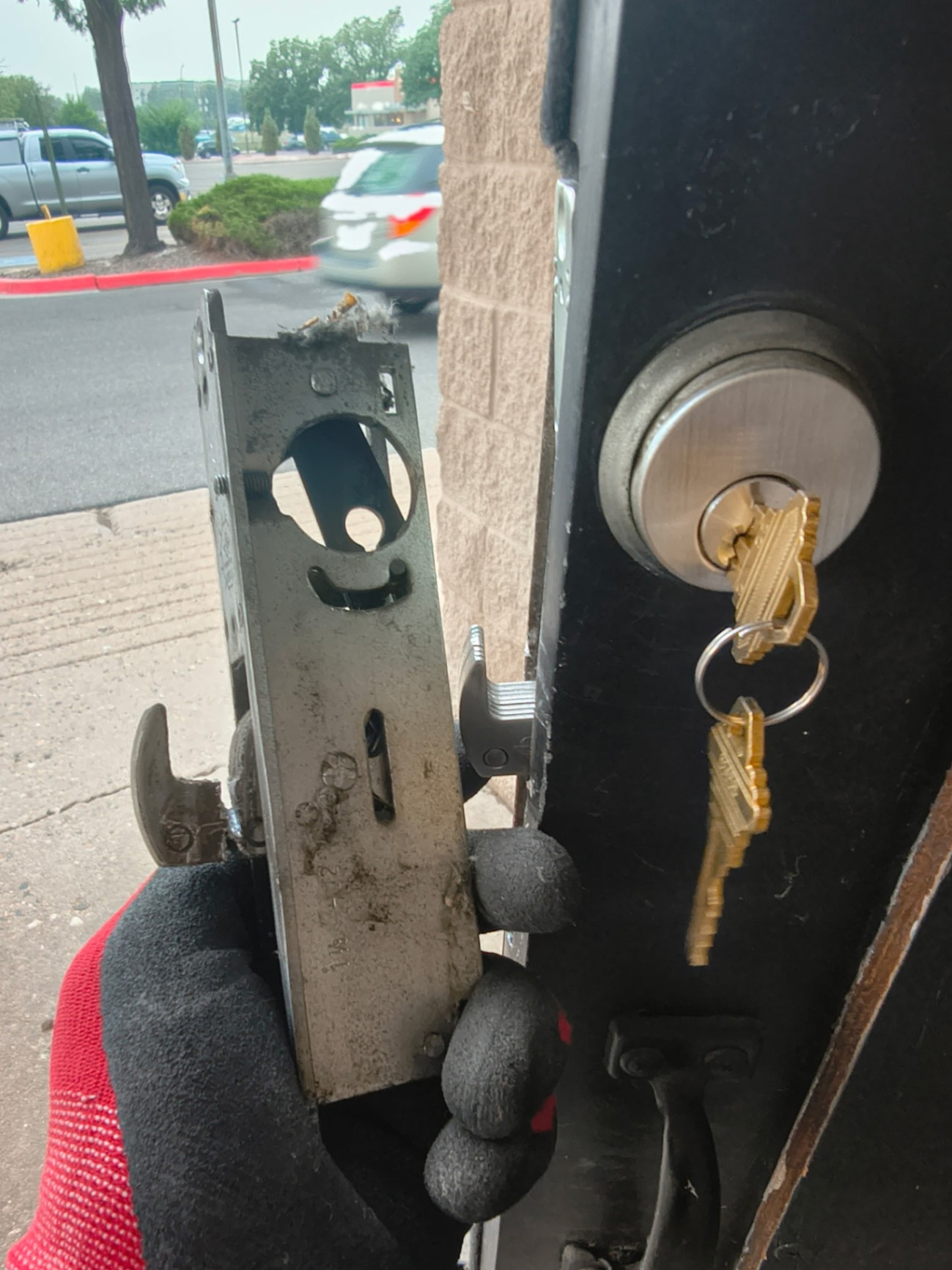 Gloved locksmith holds a removed mortise lock assembly beside a commercial door with a keyed cylinder and keys visible.