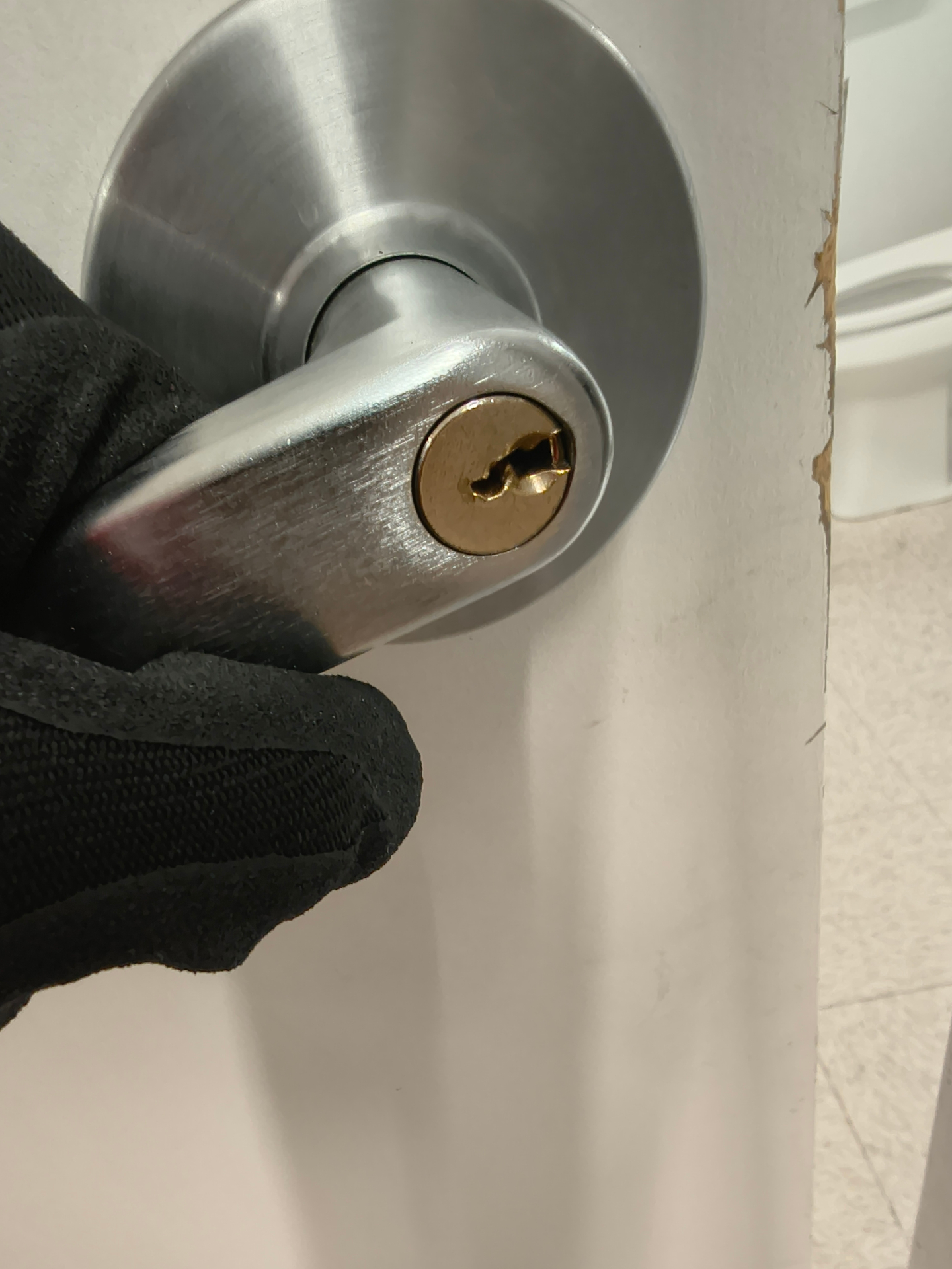Gloved locksmith holding a gold-colored cylinder and keys beside a silver commercial restroom door knob - view 3