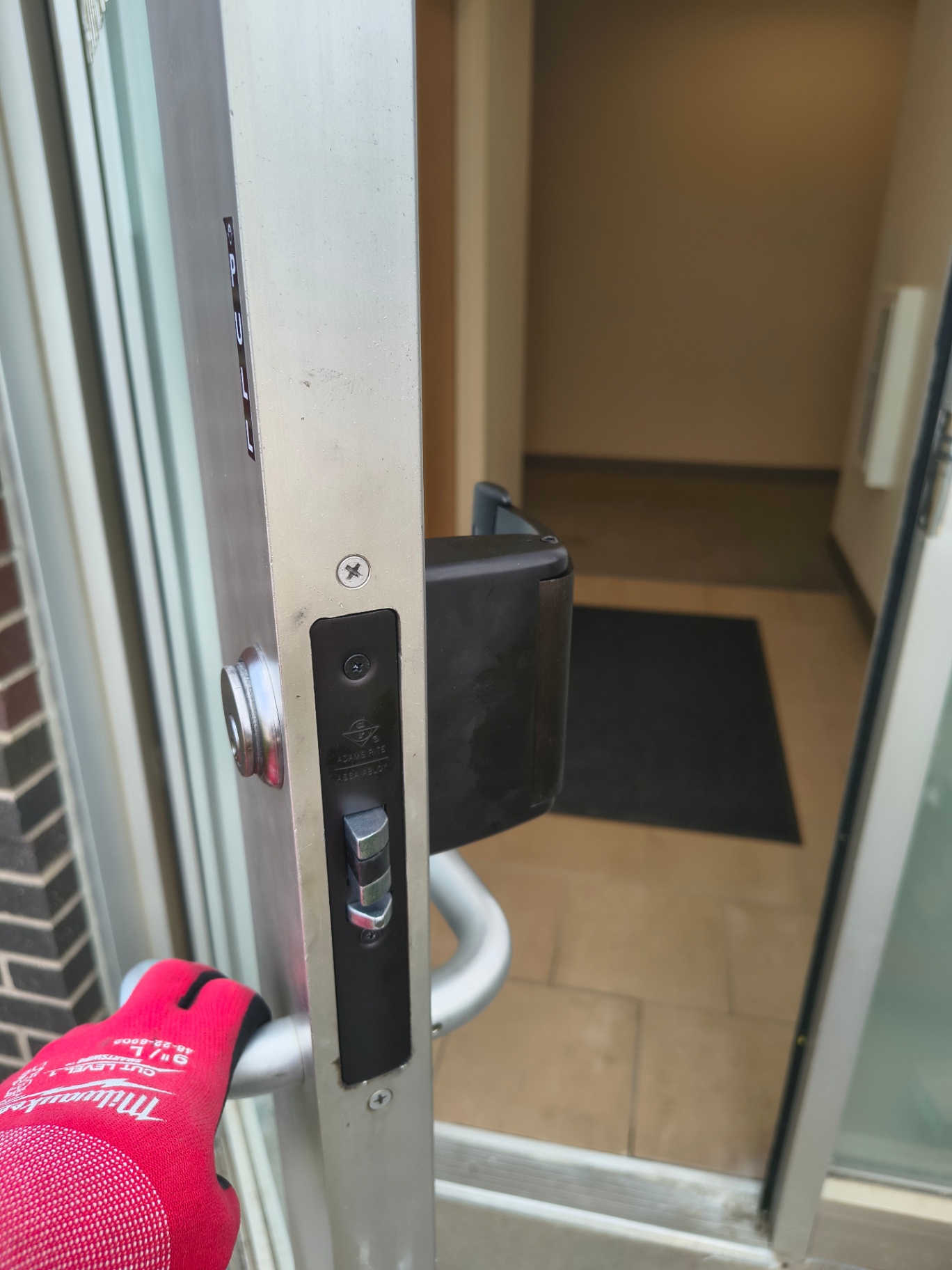 Open glass-and-aluminum commercial door showing exposed mortise lock hardware, pull handle, and black push exit device - view 4