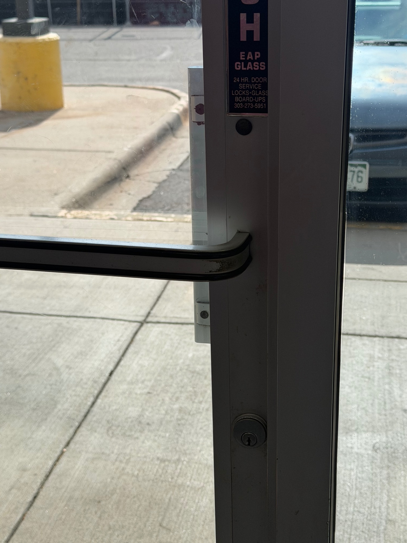 Close-up of a worn keyed cylindrical lock and pull handle on an aluminum-framed glass storefront door in Denver.