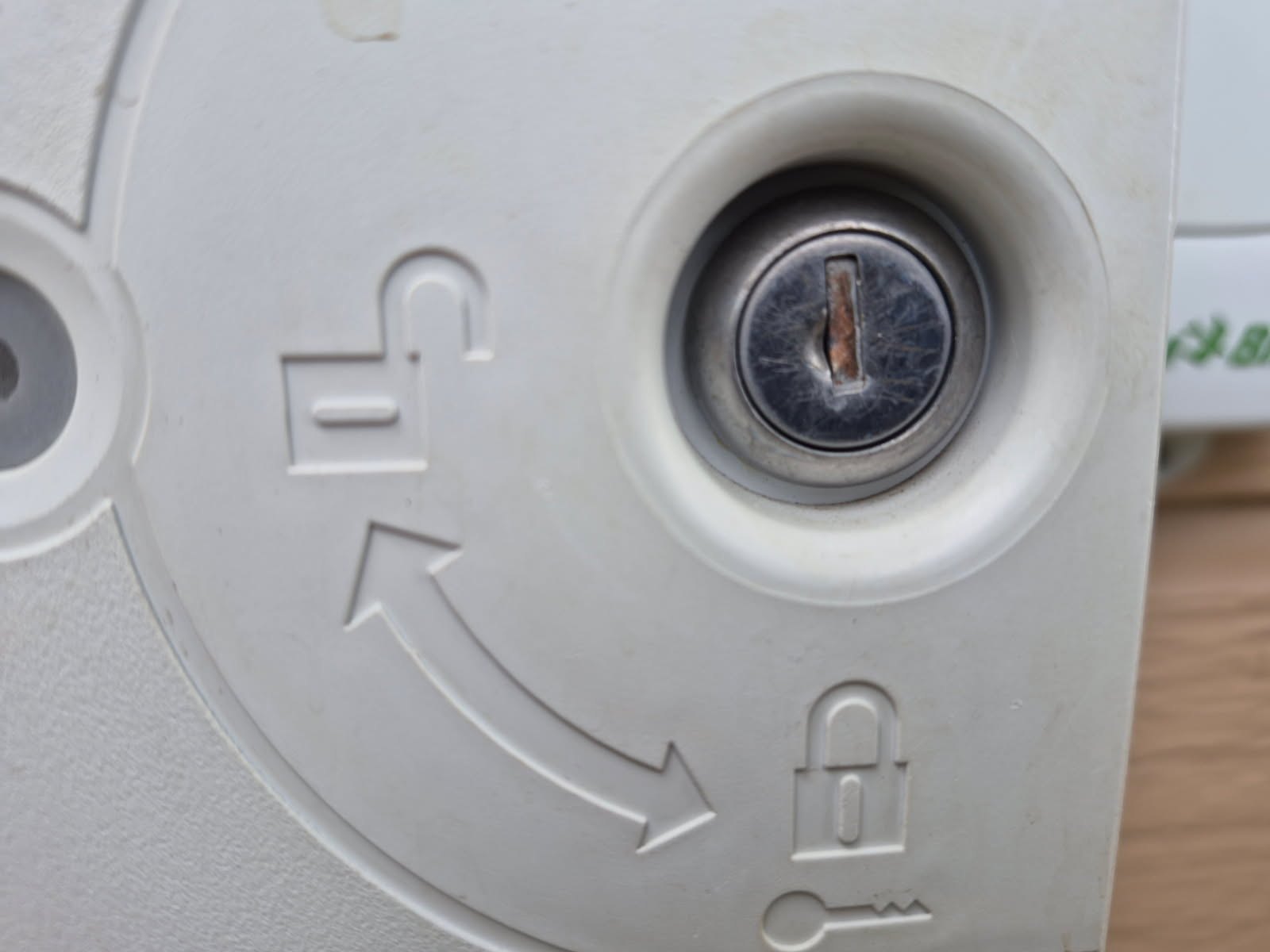 Close-up of a scratched round metal keyed cylinder in a white sprinkler-controller housing with lock and unlock arrows.